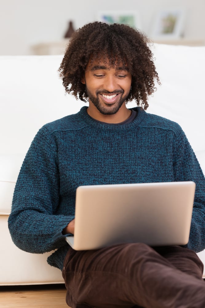 Handsome hip young African American man smiling as he types on his laptop computer while relaxing on the floor in the living room-1